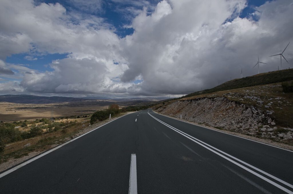 Asphaltierte Straße führt durch eine weitläufige, steinige Berglandschaft bei Livno. Rechts stehen Windkraftanlagen auf einem Höhenzug, darüber türmen sich mächtige Wolken.