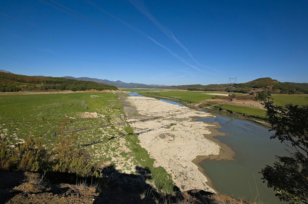 Beschädigte Hängebrücke über dem Mat bei Bushkash in Albanien, umgeben von ausgedehnten Kiesflächen, grünen Ufern und bewaldeten Bergen.