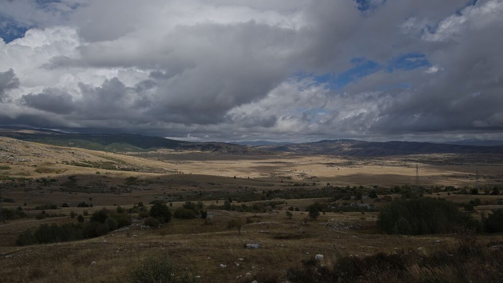 Panoramablick über eine ausgedehnte, trockene Hochebene mit vereinzelten Sträuchern, sanften Bergzügen und tief hängenden, dunklen Wolken bei Livno.