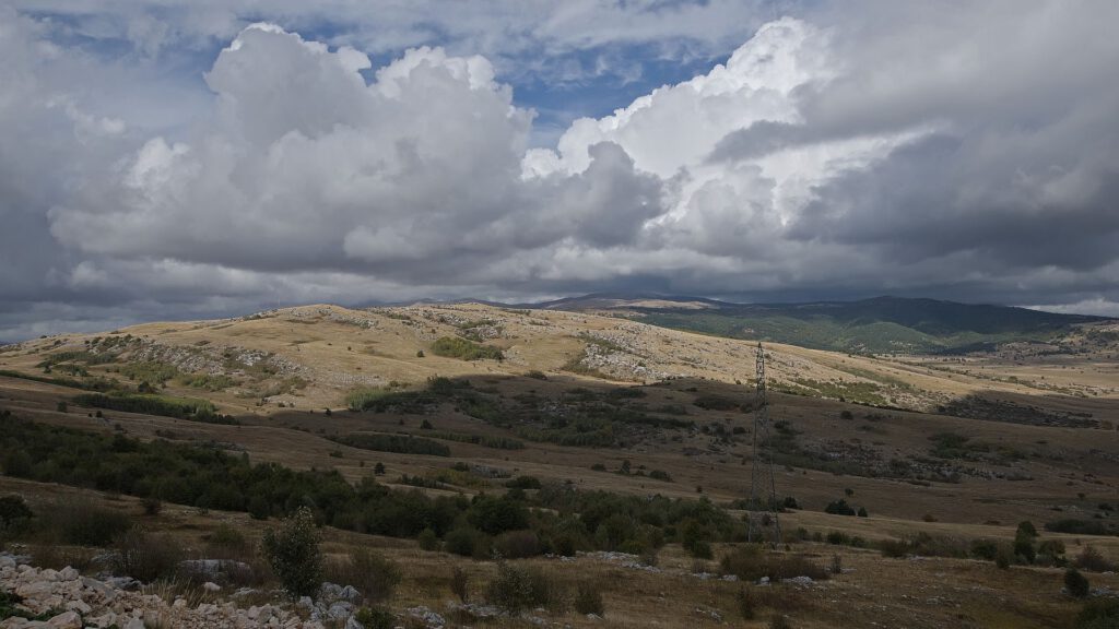 Sonnenbeschienene, steinige Hügel mit trockenen Grasflächen und vereinzelten Büschen unter mächtigen Quellwolken. Im Vordergrund steht ein Strommast.