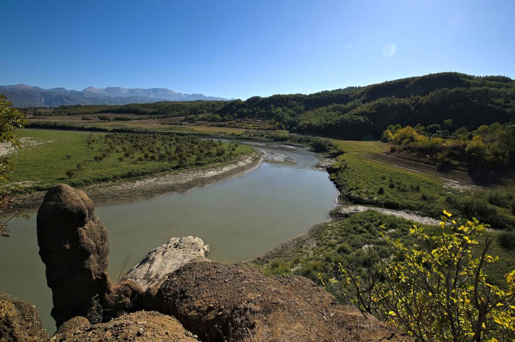 Der Fluss Mat schlängelt sich bei Bushkash durch eine grüne Talebene, eingerahmt von bewaldeten Hügeln und einem Gebirge am Horizont.