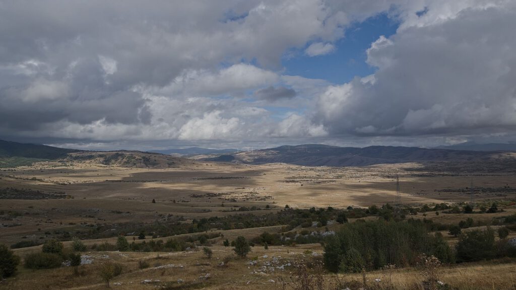 Weite, goldbraune Hochebene mit sanften Hügeln und vereinzelten Büschen. Sonnenlicht fällt durch Lücken einer mächtigen Wolkendecke auf die Landschaft.