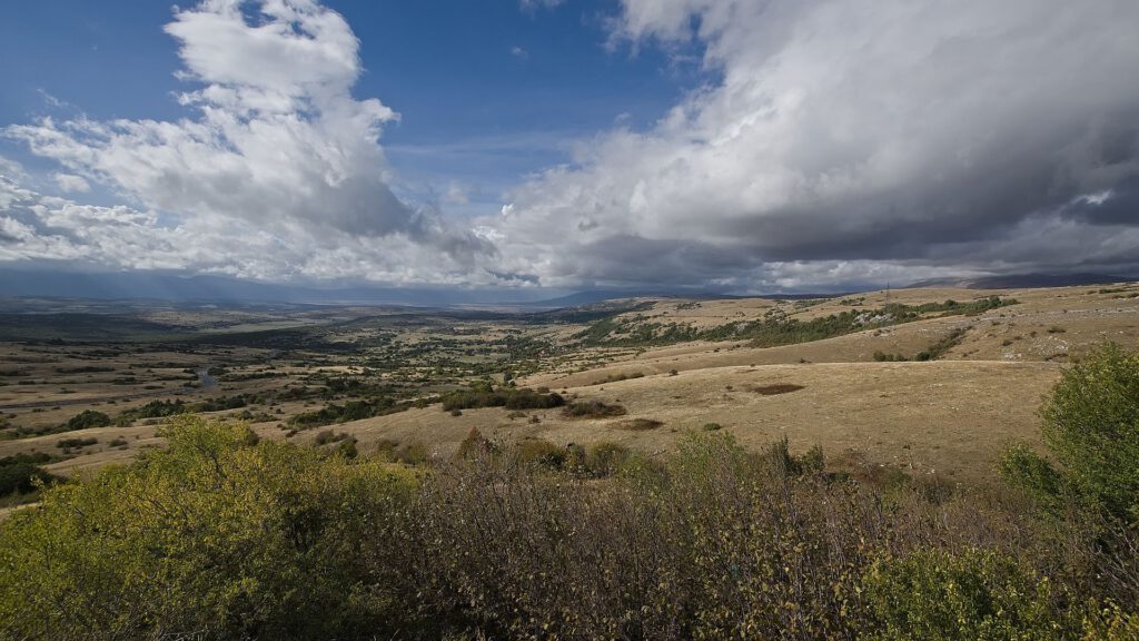 Panorama einer weitläufigen, goldbraunen Berglandschaft mit vereinzelten Sträuchern und einer kurvigen Straße links im Tal. Darüber wechseln sich blauer Himmel und große Wolkenfelder ab.